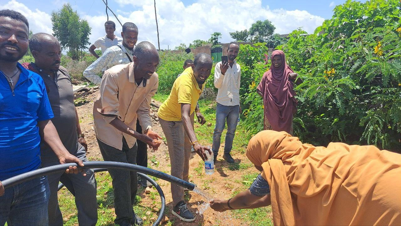 Community members smile and watch as a woman in a yellow headscarf stretches out to wash her hands with clean water flowing from a hose.