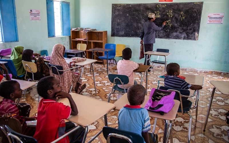 Students sit at desks in a classroom while an instructor writes on a chalkboard.