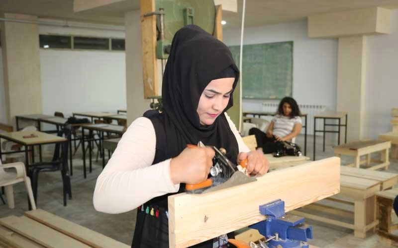 A student in a workshop classroom uses a hand plane to shape a piece of wood secured in a vise, with worktables and another student in the background.