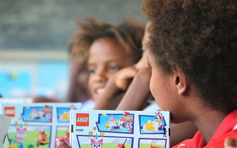Children sit together around educational building-block materials in a classroom, with a box of activity sets visible in the foreground.
