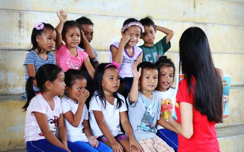 Children sit on benches in a classroom while an instructor leads a lesson, with several children raising their hands.