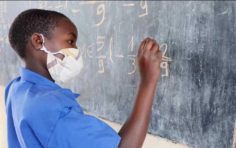 A student wearing a face mask writes math equations on a chalkboard during a classroom lesson.