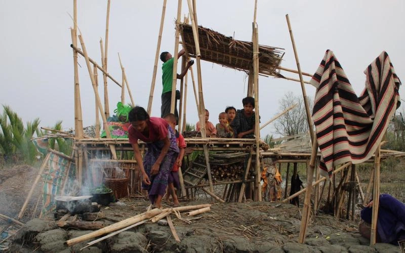 A man and woman work to reassemble their destroyed bamboo dwelling, with multiple children looking on.
