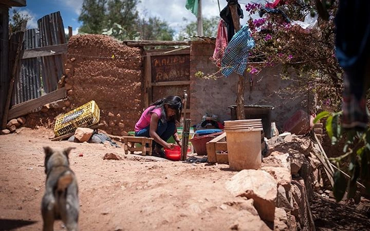 A girl or young woman crouches outside a hut made of mud, washing a red bowl, while a dog appears to be walking toward her.