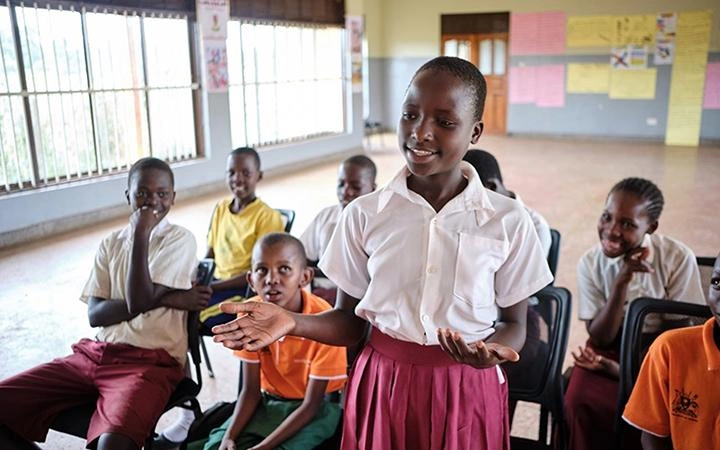 An adolescent girl stands in front of a small group of seated boys and girls, facing toward the camera.