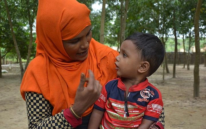 A small boy sits on the lap of a woman wearing a bright orange headscarf, looking toward her face as she speaks to him.