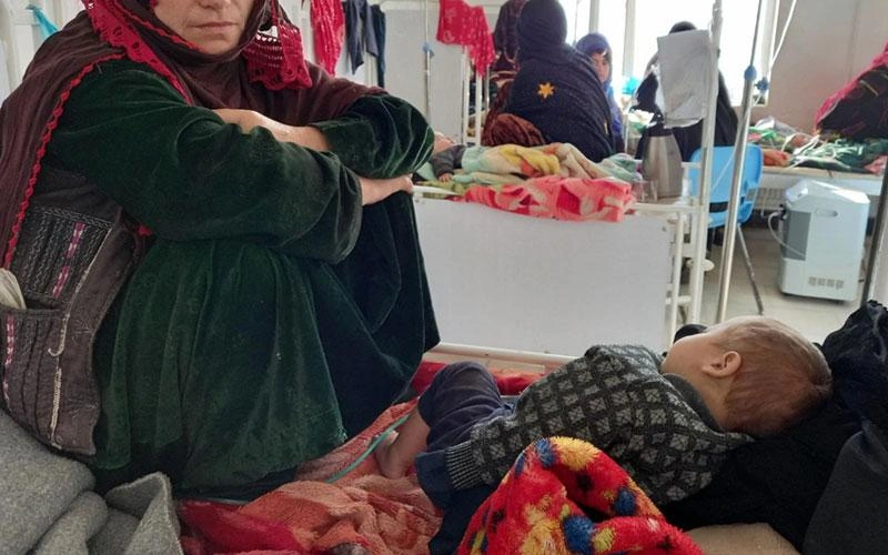 A toddler or young child lies listlessly on a bed while a mother sits beside.