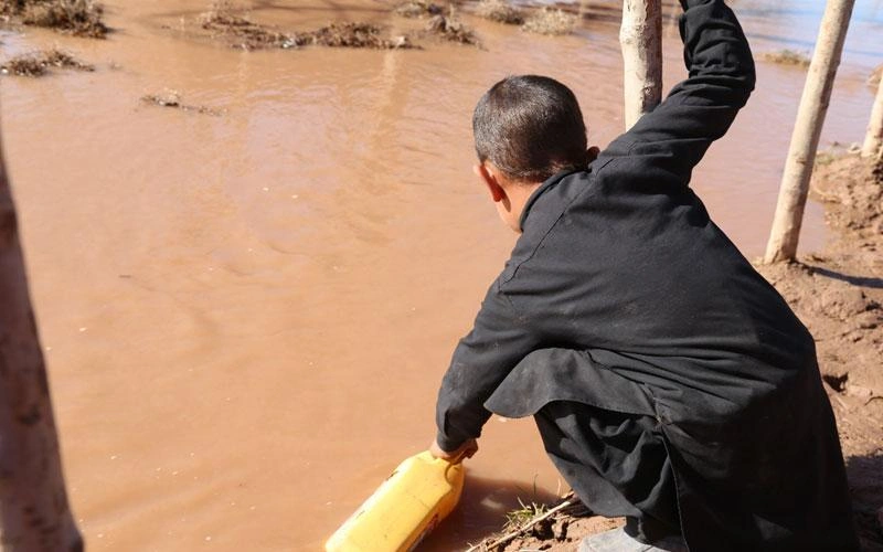 A child crouches by a large, muddy puddle, scooping water in a yellow jug.