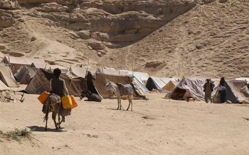 A group of tents on the dry sand at the foot of a rocky mountain. In the foreground, a man carries jugs of water on a donkey.
