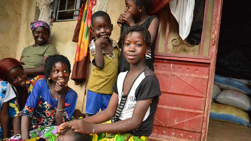 A group of children gather together in front of a doorway, with Alimatou braiding another girl’s hair and an adult woman sitting in the background.