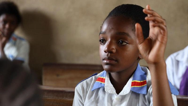 Blessing raises her hand in her classroom, sitting among other school children.