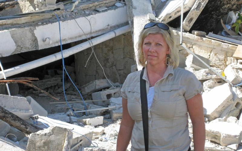 A Canadian woman looks at the camera, rubble from collapsed buildings behind her.