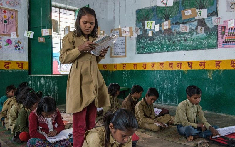 A young Indian girl stands in her classroom and reads aloud from the book she holds in her hand.