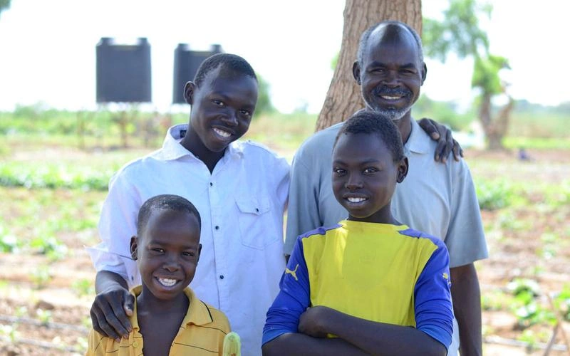 A family from Sudan - dad and three young sons - stand in the shade of tree. There is garden field with water tanks in the background.