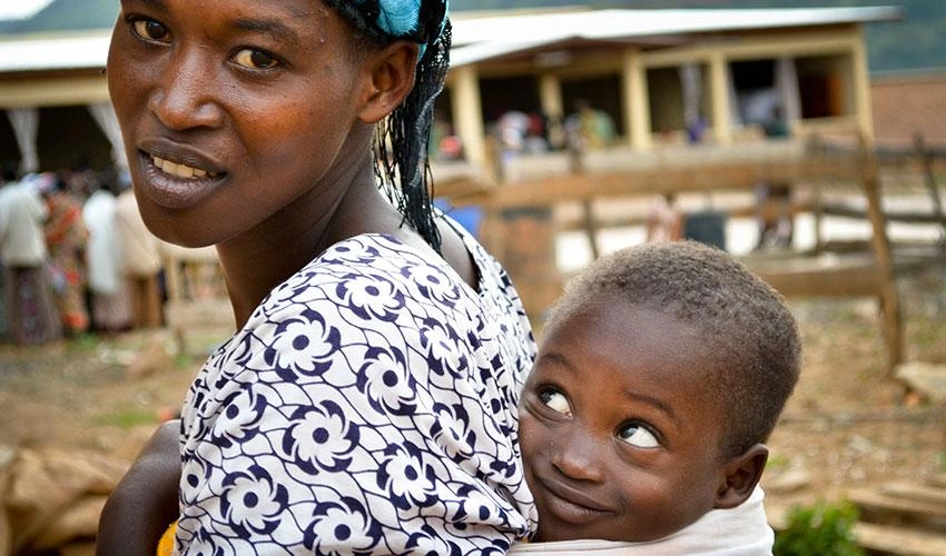 A mom carries her young son in a wrap attached to her back, and he looks up at her admiringly.