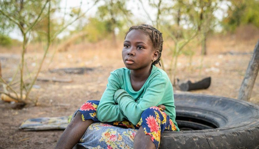 Child in a green shirt, sitting on a tire in a rural area. (Ghana, 2025)
