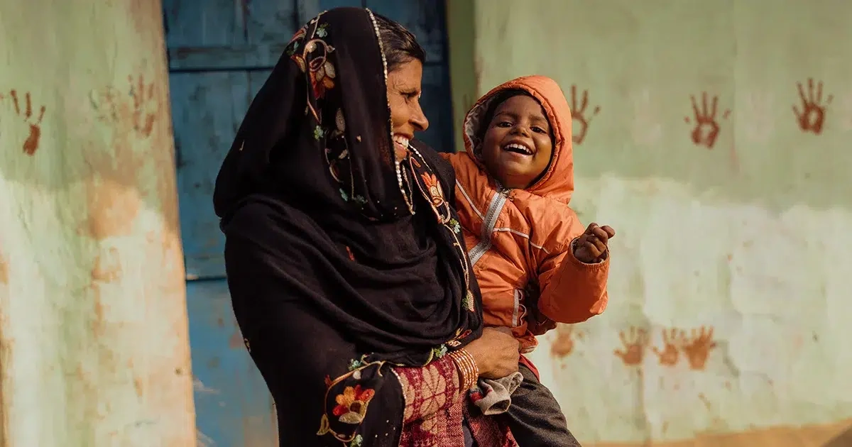 A woman holds a small child outside a home with painted handprints on the wall behind them.