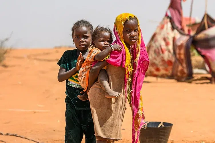 Three Sudanese children stand together on sandy ground in a spontaneous settlement at Adre, Chad.