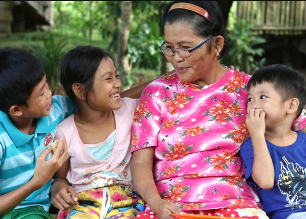 Grandmothers in east Asia smiles fondly at her grandchildren as they sit outdoors in the shade.