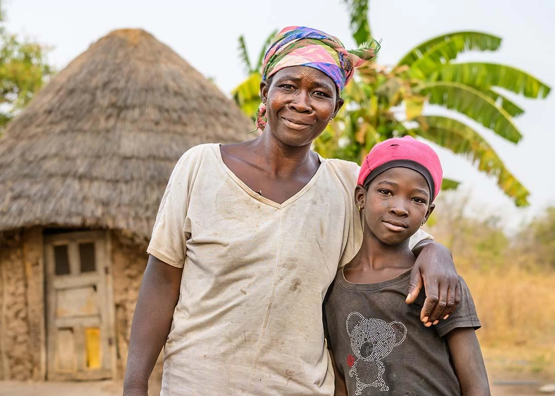 Smiling woman has arm around young girl in from of a clay outise in fron of a small hut with thatched roof in Africa.