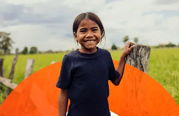 A young girl smiles, standing outdoors, resting her left hand on a wooden fence post.