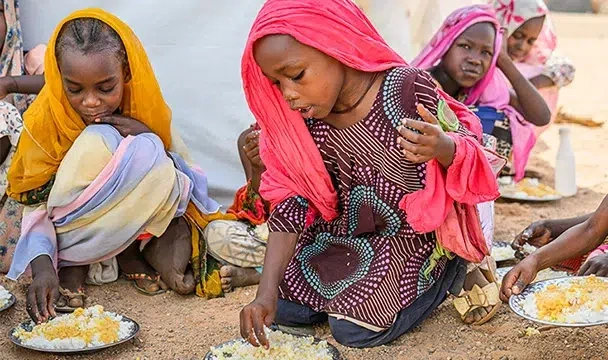 A group of girls wearing headscarves sitting on the ground in a Sudanese refugee camp and eating from plates of donated food.