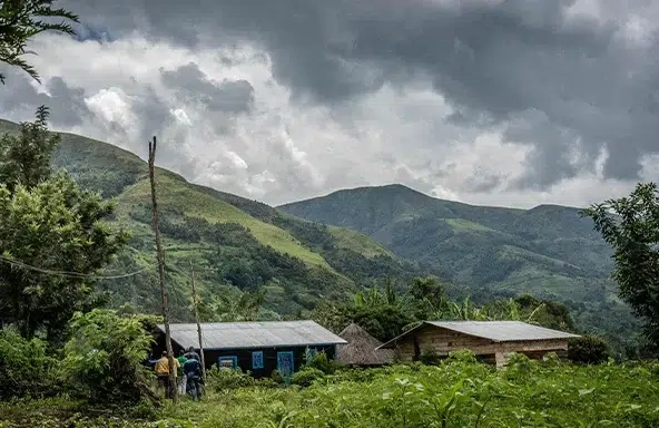 A wide shot image of a green forested area in the Democratic Republic of Congo. In the distance there are people walking towards two large cabin’s.