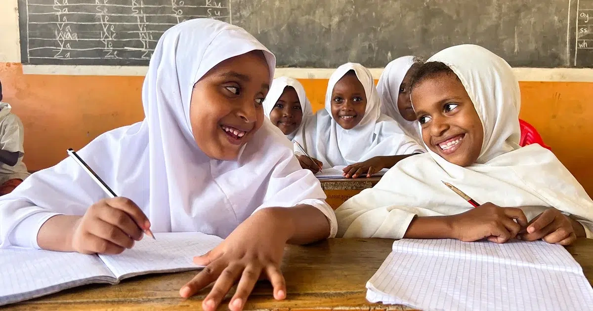 Two students are smiling at each other while sitting at a desk inside a classroom.