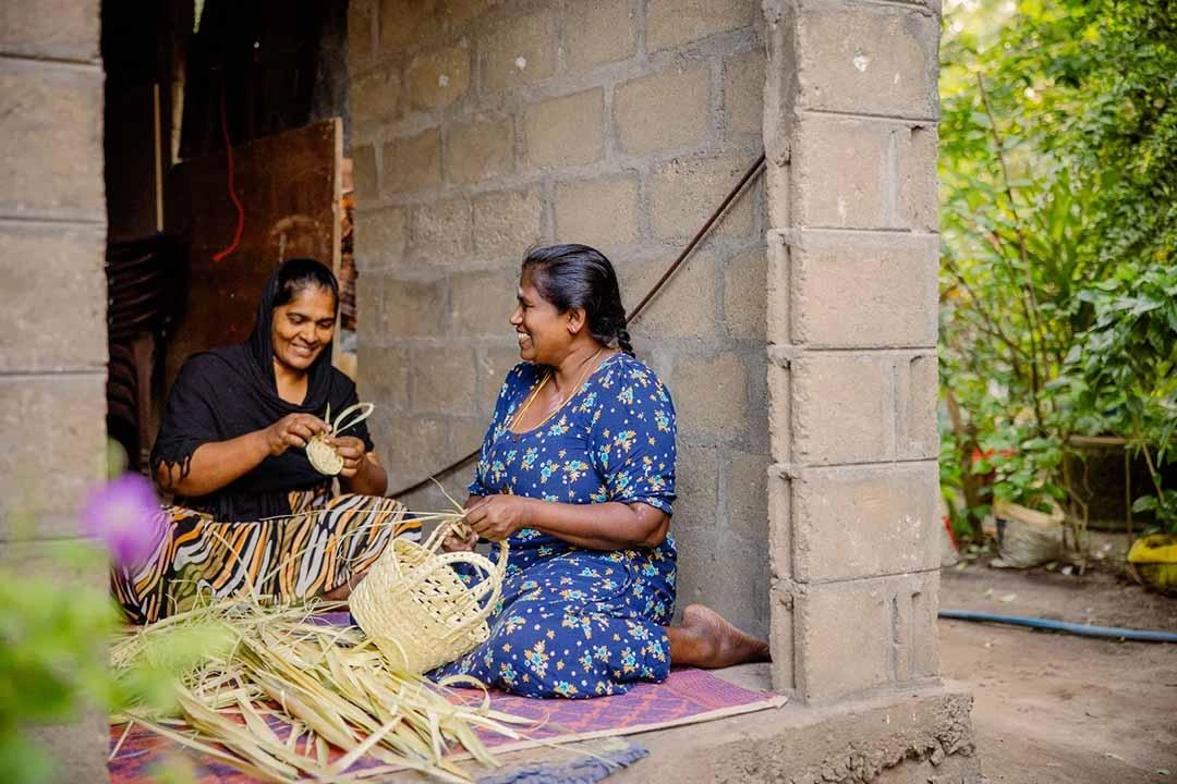 Women sit and smile while making baskets to sell.