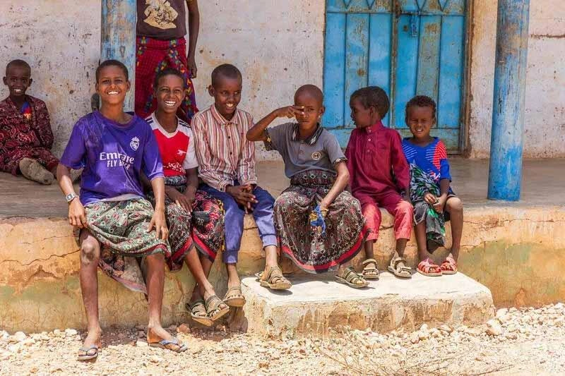 Refugee children sit and smile in a dusty rural area.