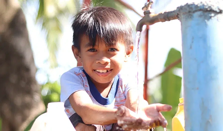 A little boy smiles as he washes his hands at an outdoor faucet.