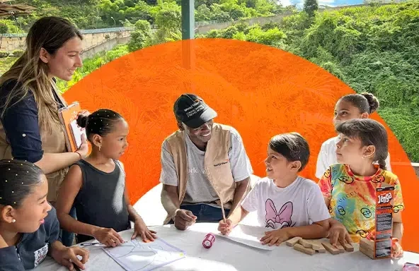 orld Vision staff leading outdoor educational activity with children, featuring drawing materials and games on a table in a lush, green setting.