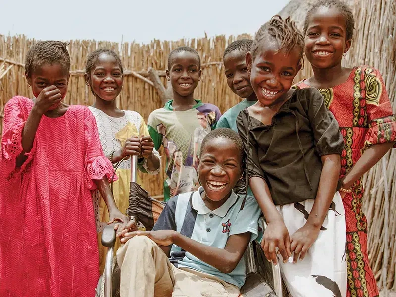 Group of children standing and smiling together around a child in a wheelchair in a rural village setting with woven reed walls in the background.