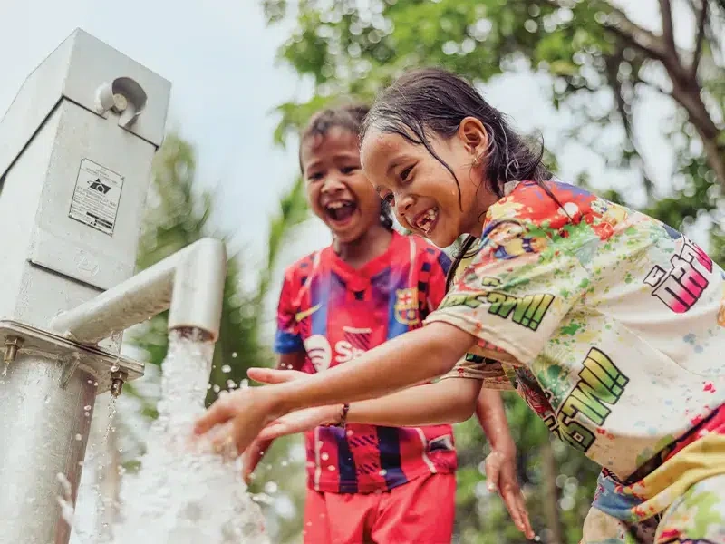 Two children reaching out to wash their hands as fresh water flows out from a hand pump.