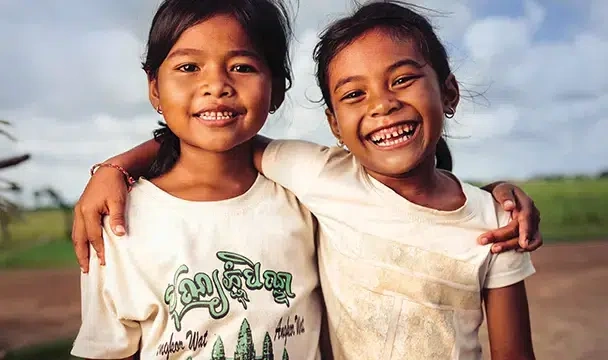 Two young girls wearing white t-shirts smile broadly as they stand side by side in a grassy field, their arms on each other’s shoulders. (Cambodia, 2025)
