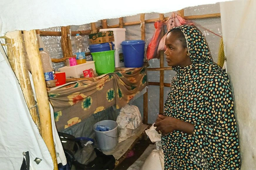 A female refugee stands in the cooking area of her temporary shelter. A mix of plastic bottles and buckets contain limited resources for food and survival.