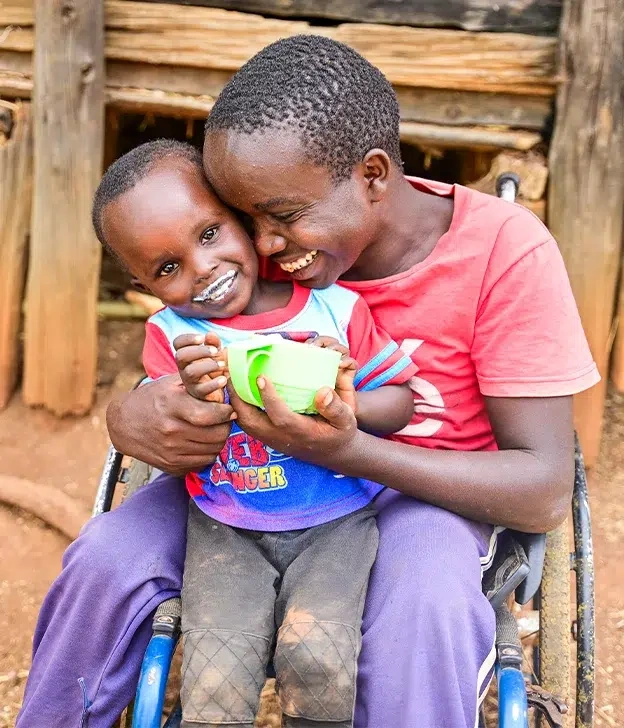 A woman sitting in a wheelchair hugs a young child while sharing a cup outside a home.