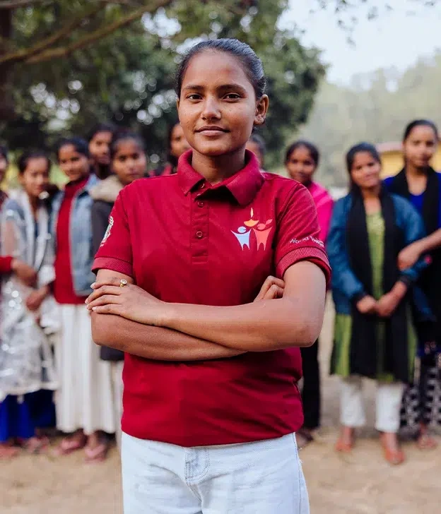 A young community leader stands with arms crossed in front of a group of peers during an outdoor gathering.