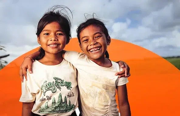 Two young girls wearing white t-shirts smile broadly as they stand side by side in a grassy field, their arms on each other’s shoulders. (Cambodia, 2025)