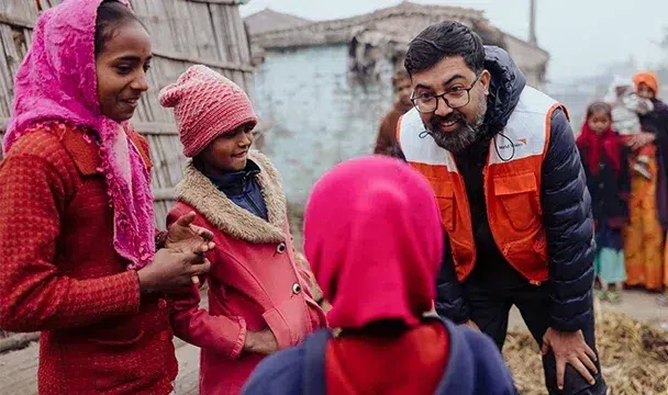 A World Vision staff in an orange safety vest speaking with children in a residential community.