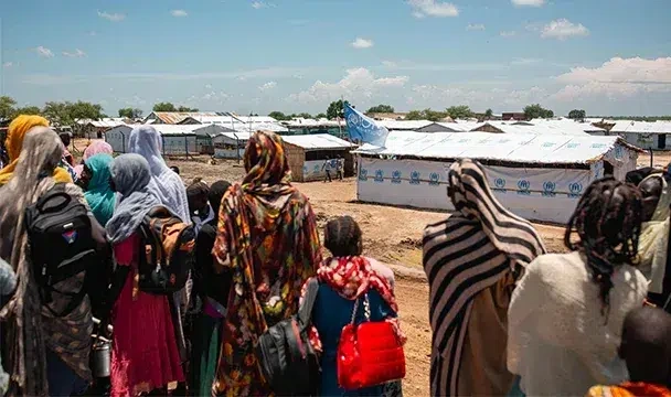 Group of people standing near a camp of white temporary shelters under a clear sky.