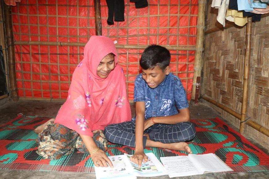 Un enfant lit un livre en compagnie d’un adulte dans un abri rudimentaire. (Bangladesh, 2024)