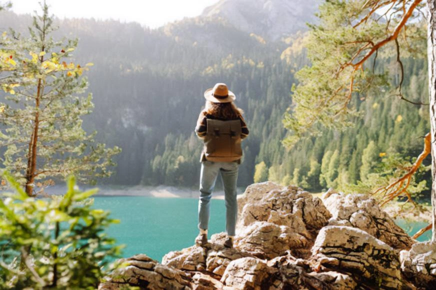Une femme debout sur des rochers, portant un sac à dos et un chapeau Tilly, contemplant un lac paisible entouré d’arbres.