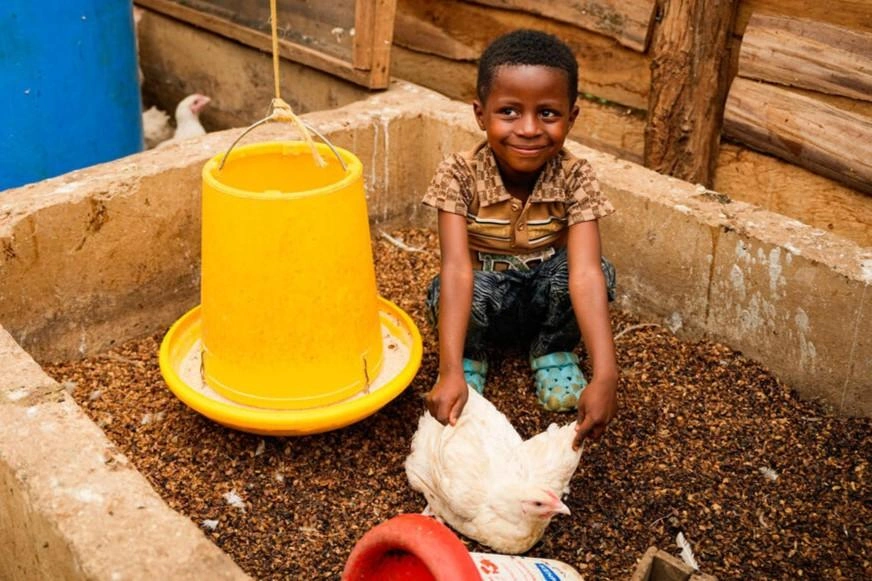 Un garçon d’âge primaire est assis, recroquevillé à côté d’une poule, et sourit.