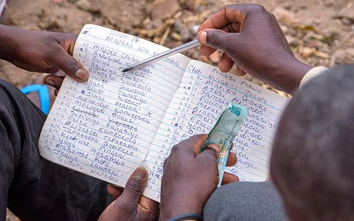 Une paire de mains tient un registre dans lequel sont inscrites des colonnes de noms. Une autre paire de mains tient de l’argent, tout en pointant la liste avec un stylo.