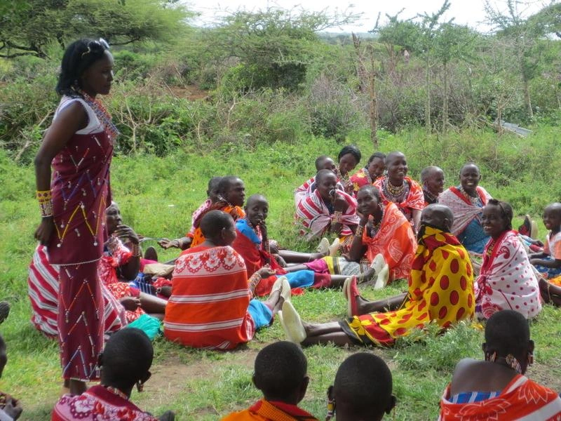 Un groupe de femmes vêtues de robes aux couleurs vives est assis sur l'herbe. Une femme se tient debout parmi elles.
