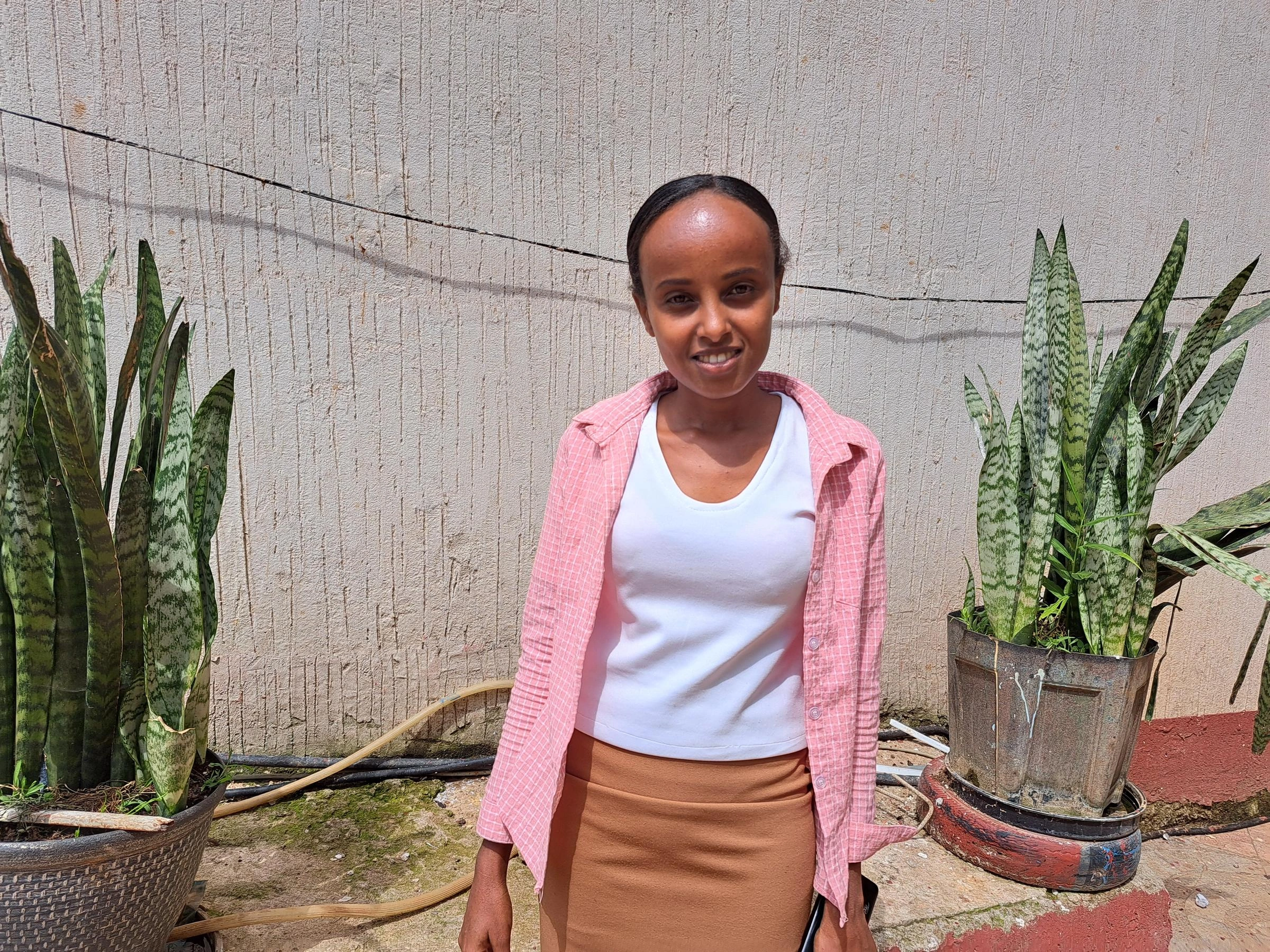 Une jeune femme vêtue d’un chandail rose et blanc se tient devant deux plantes en pot et sourit.