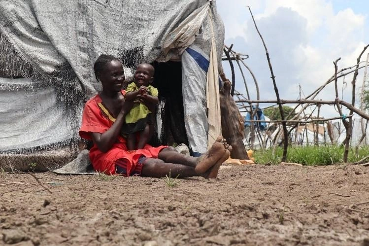 Une femme est assise devant une tente, souriante, son enfant dans les bras.
