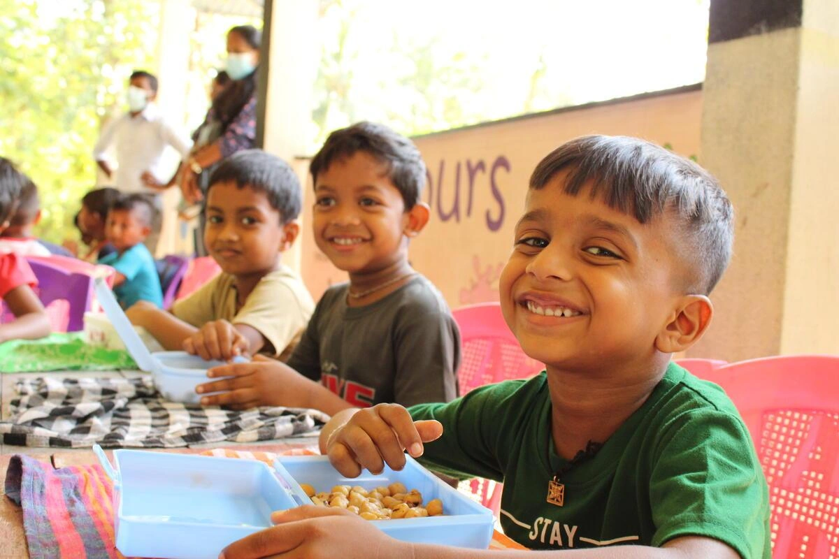 Des enfants sourient en dégustant leur dîner dans des contenants bleus, assis à une longue table.