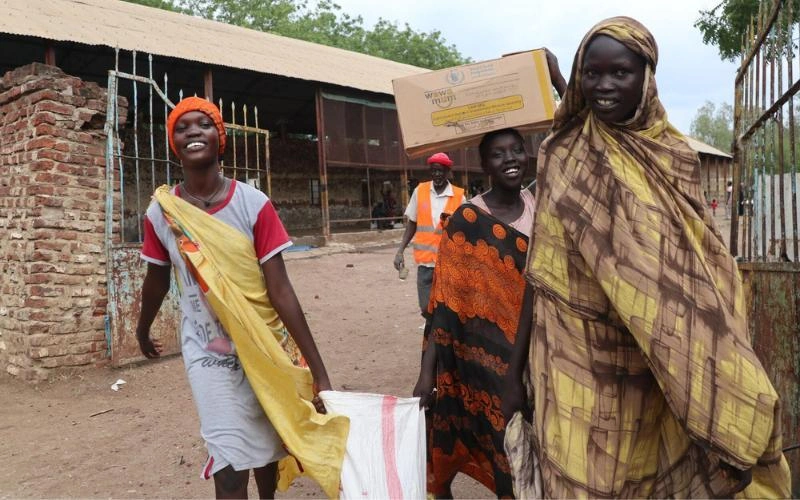 Trois femmes souriantes sortent d’une enceinte à Malakal, au Soudan du Sud, en transportant des sacs et des boîtes de nourriture.